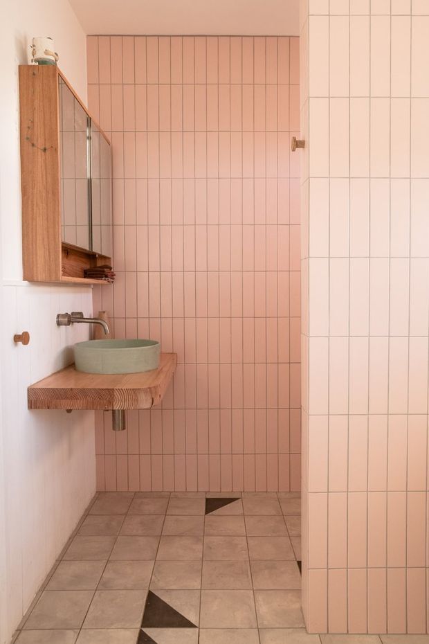 A compact contemporary bathroom with a walk-in shower, vertical blush-pink tiled feature wall, pale square floor tiles, and a partial tiled partition. A timber-framed mirrored cabinet and thick solid timber vanity shelf support a round vessel basin with a wall-mounted metal tap. The space has a warm, softly coordinated palette with small dark geometric floor accents.