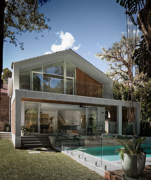 Exterior view of a contemporary multi-level residence set within a leafy garden. The house features a strong horizontal concrete frame, extensive floor-to-ceiling glazing, timber wall cladding, a sloping roof, and a glass-balustraded upper terrace. A turquoise swimming pool occupies the foreground beside the lower-level living spaces.
