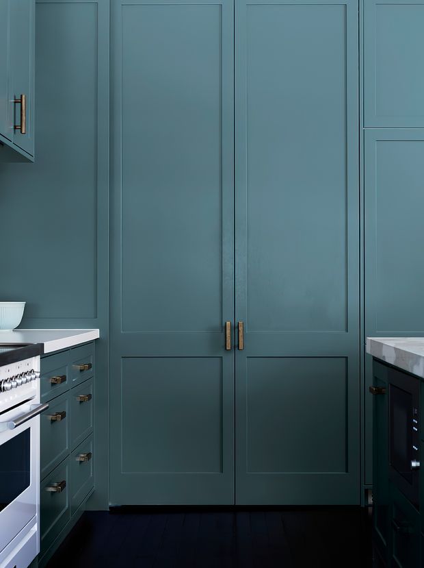 A compact, richly coloured kitchen centred on tall blue-green shaker-style cabinetry with recessed panel doors and brass handles. Matching lower cabinets flank a central double-door pantry or refrigerator enclosure, with pale stone benchtops, dark timber flooring, and partially visible integrated appliances.
