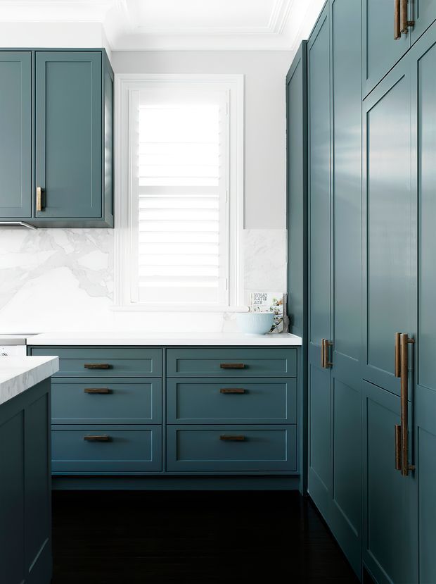 A compact, refined kitchen with deep blue-green shaker-style cabinetry, tall pantry cupboards, brass hardware, white marble-look splashback and benchtops, dark timber flooring, and a bright shuttered window. The cabinetry and detailed trim create a classic, tailored interior.