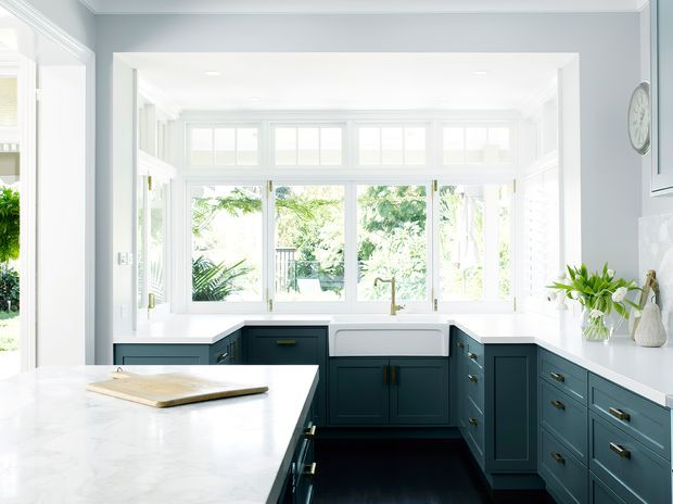 Bright, refined kitchen with deep blue-green shaker cabinetry, white marble-look benchtops, a large central island, farmhouse-style sink, and extensive white-framed windows overlooking greenery. Pale grey walls and white trim create an airy, classic interior.
