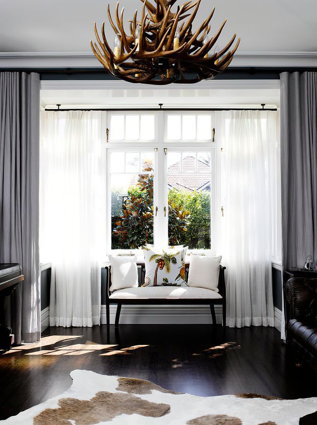Bright, refined living room centered on a tall multi-pane window with sheer white curtains and grey drapes. A black-framed upholstered bench sits beneath the window, while a dark leather sofa, polished dark timber floor, cowhide rug, and prominent antler chandelier create a classic country-inspired interior with strong natural light.