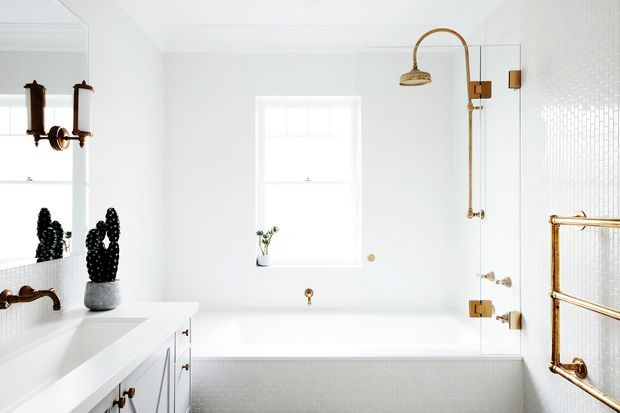 Bright, refined white bathroom featuring a built-in bathtub beneath a window, a glass-enclosed shower with warm brass fixtures, a white vanity with dark hardware, and a brass towel rail. White textured wall and tub finishes create a clean, high-contrast look with black and brass accents.