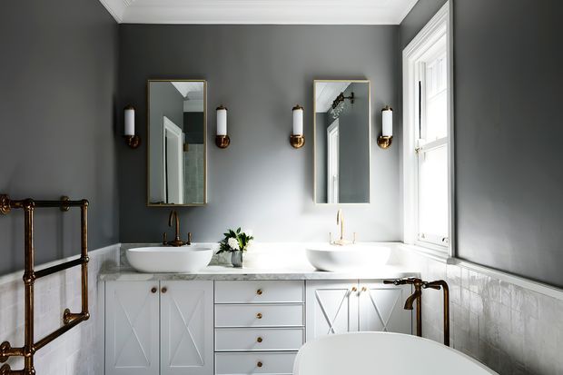 Elegant traditional bathroom with a long white double vanity, twin vessel basins, brass fixtures, framed mirrors, wall sconces, marble-look wall tiling, dark grey painted walls, and a freestanding bathtub in the foreground. White trim and a large window provide natural light.