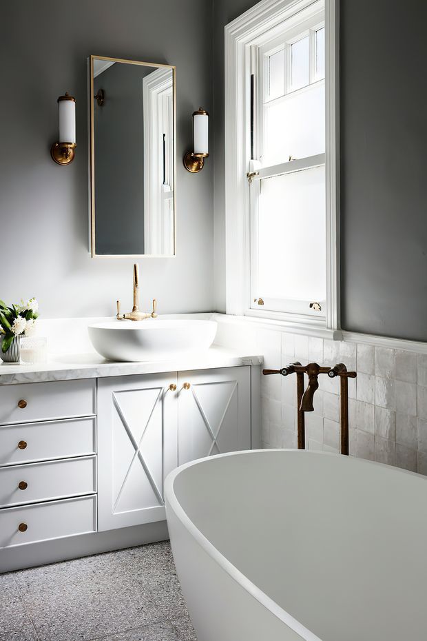 A refined traditional bathroom with a white freestanding bathtub, white vanity with cross-panel cabinetry, vessel sink, marble-look countertop, tiled lower walls, and a large white-trimmed sash window. Dark grey walls, brass wall sconces, aged brass plumbing fixtures, and a rectangular mirror add contrast and classic character.