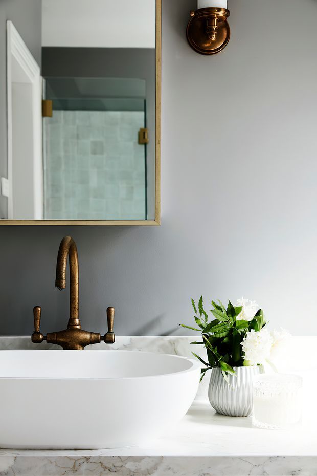 Close-up of a refined bathroom vanity with a white vessel basin on a white marble countertop, an aged brass bridge-style faucet, grey painted wall, brass-framed mirror, and matching brass wall sconce. A glass shower enclosure with pale tiled walls is reflected in the mirror, while a white flower arrangement adds a decorative accent.