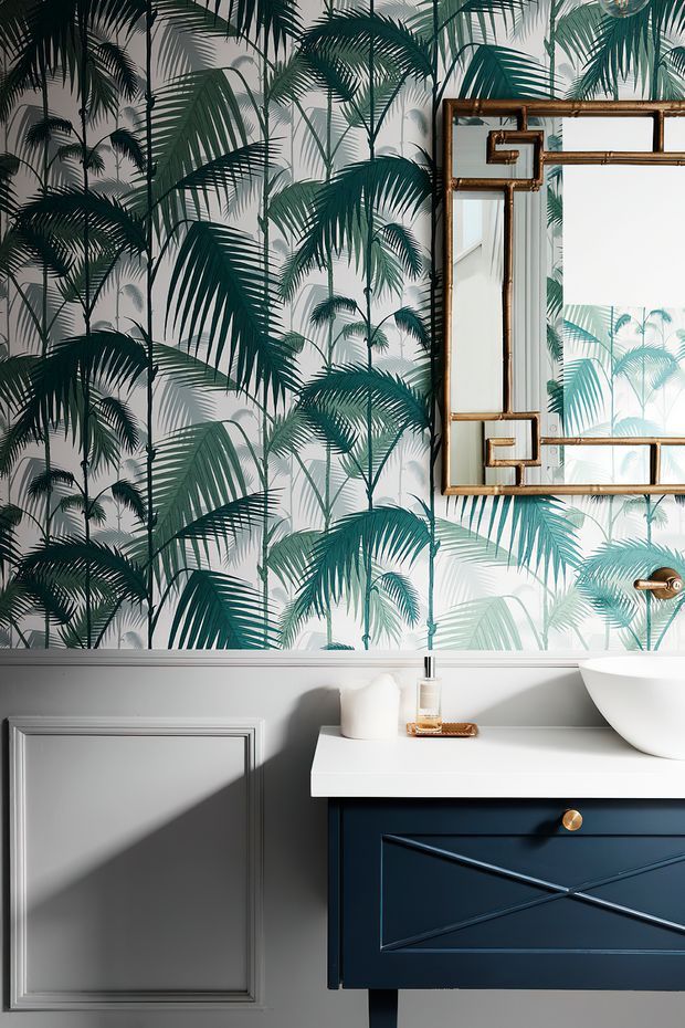A stylish bathroom vanity vignette featuring a dark navy-blue built-in cabinet with geometric panelled doors, a white stone benchtop, and a white vessel basin. The lower wall is painted light grey with decorative rectangular wall moulding, while the upper wall is covered in bold tropical palm-leaf wallpaper in deep green and turquoise tones. A large antiqued brass-framed mirror and brass tapware add a refined vintage accent.