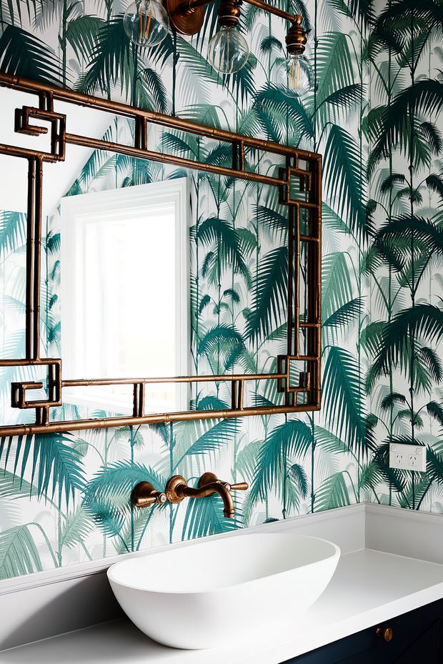 A stylish bathroom featuring a freestanding white oval bathtub on a dark vanity or platform, a wall-mounted copper-toned bath filler, and a large rectangular mirror with an industrial pipe-style frame. The walls are covered in bold tropical palm-leaf wallpaper, complemented by exposed copper-toned pendant light fittings and white trim.