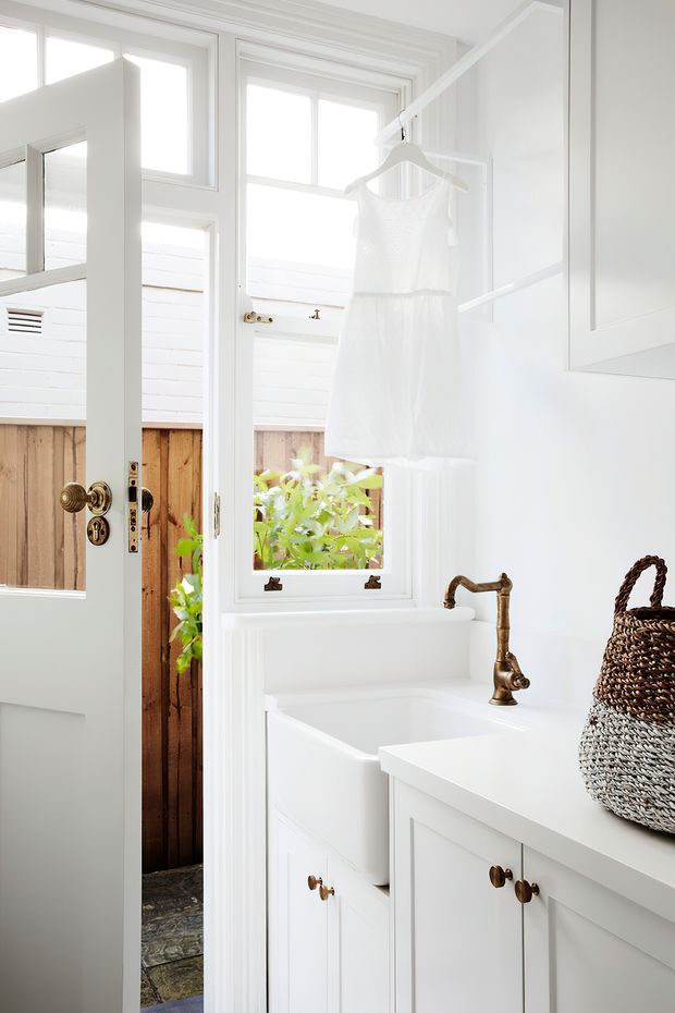 Bright white bathroom with traditional panelled cabinetry, a freestanding bathtub beneath large windows, and a bronze or aged brass tap. A partially open glazed door reveals a timber-fenced outdoor area. The room has white-painted trim and walls, stone-look flooring, and a woven basket on the vanity.
