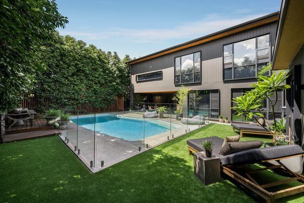 A contemporary two-storey dark-clad residence arranged around a landscaped backyard pool. The image shows a glass pool fence, turquoise swimming pool, artificial lawn, timber decking, outdoor lounge furniture, dense screening vegetation, and large glazed openings connecting the house to the outdoor area.