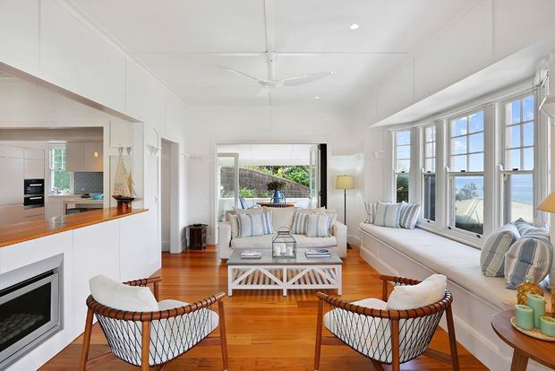 Bright coastal-style living room with warm timber flooring, white paneled walls and ceiling, a large built-in window seat, white sofas, and two woven lounge chairs. The room opens toward a kitchen and a glazed outdoor-facing area, with ocean or water visible beyond the windows.