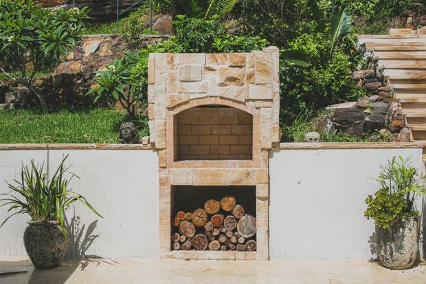 Outdoor masonry fireplace built into a white rendered retaining wall, with a stone-clad surround, arched firebox, and lower wood storage niche filled with log sections. Tropical landscaped planting, stone retaining walls, and paved flooring surround the feature.