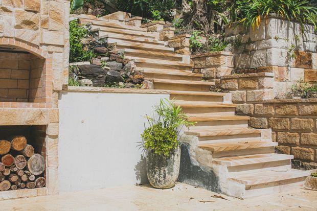 Sunlit outdoor stone stairway ascending through a landscaped garden, with warm sandstone retaining walls, a built-in arched wood-storage niche, natural rock planting beds, and a potted plant. The scene has a rustic Mediterranean feel and warm earthy tones.