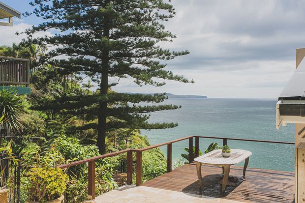 A timber outdoor deck overlooking the ocean, surrounded by lush coastal vegetation and mature Norfolk pine trees. The deck has a simple outdoor table and chairs, dark timber balustrading, and a partial view of adjacent residential structures. The scene is bright and expansive with a relaxed coastal outlook.