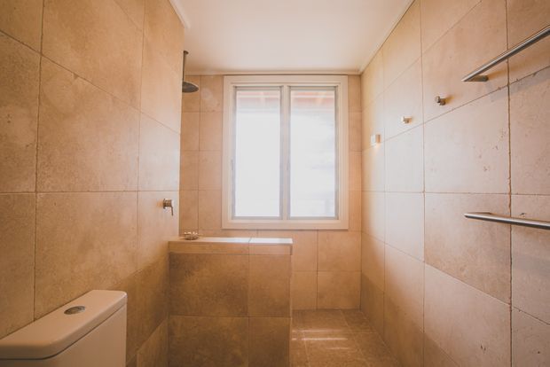 A compact, warm-toned tiled bathroom with a walk-in shower area, freestanding-style tiled bathtub beneath a frosted window, toilet partially visible at left, and wall-mounted chrome towel rails on the right. Large beige stone-look tiles cover the walls and floor, creating a simple contemporary space with natural light.