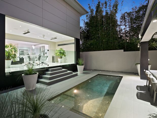 Contemporary residential courtyard at dusk featuring a compact rectangular swimming pool, broad pale tiled paving, integrated steps, sculptural planters, and a dramatic black-framed glazed opening into a bright dining and living area. Tall screening vegetation and rendered boundary walls create a private outdoor setting, while recessed and concealed lighting adds a refined evening atmosphere.