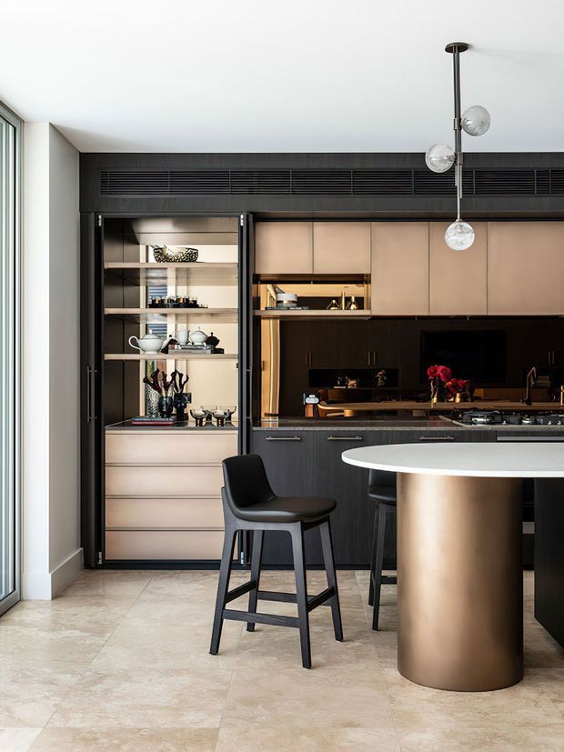 A refined contemporary kitchen with a dark charcoal island and cabinetry, integrated open display shelving, warm bronze-toned upper cabinetry, and a cylindrical metallic breakfast-bar end. A black bar stool and sculptural globe pendant light hang over large pale stone-look floor tiles. Full-height glazing provides abundant natural light.