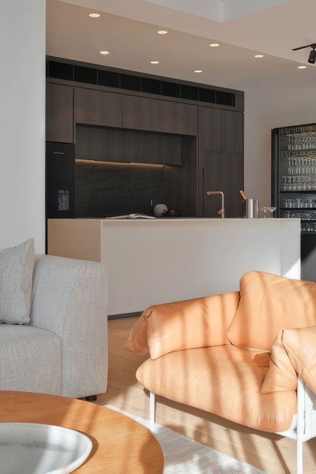 A contemporary open-plan kitchen and living area viewed from the lounge. The kitchen features dark timber-look full-height cabinetry, integrated appliances, a dark stone-like splashback, under-cabinet lighting, and a pale island with a brass-toned tap. A light grey sofa and warm tan leather lounge chairs sit around a rounded timber coffee table, with strong sunlight casting shadows across the room.