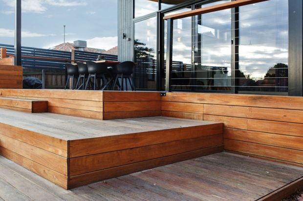 A contemporary outdoor timber deck and stepped terrace beside a glazed building facade. Warm timber decking and raised planters frame an outdoor bar or dining area with black stools, while large dark-framed sliding glass doors reflect the sky and surrounding trees.