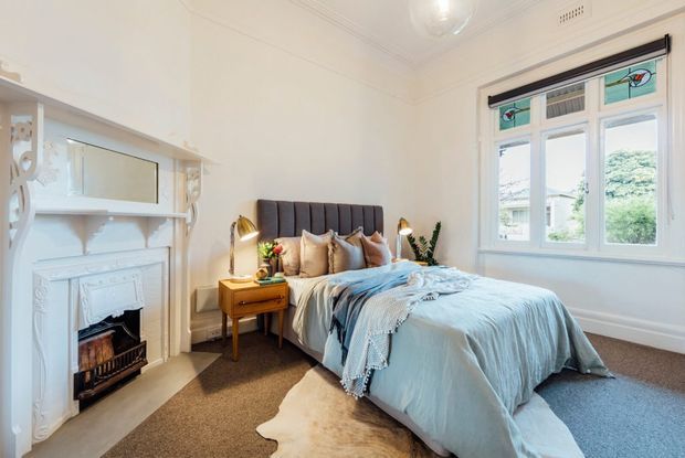 Bright, traditionally detailed bedroom with a centered upholstered bed, pale blue bedding, matching bedside tables and lamps, a decorative white fireplace and mantel, built-in shelving, decorative stained-glass window panels, and carpeted flooring. The room has high ceilings, ornate cornices and a calm neutral palette with soft blue accents.