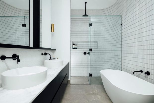 A contemporary bathroom with a freestanding white bathtub, double vanity with vessel basins, black wall-mounted tapware, and a frameless glass shower enclosure. White small-format tiled walls with fine dark grout create a curved grid effect, while pale stone-look flooring, black cabinetry, and brass wall lighting add contrast.
