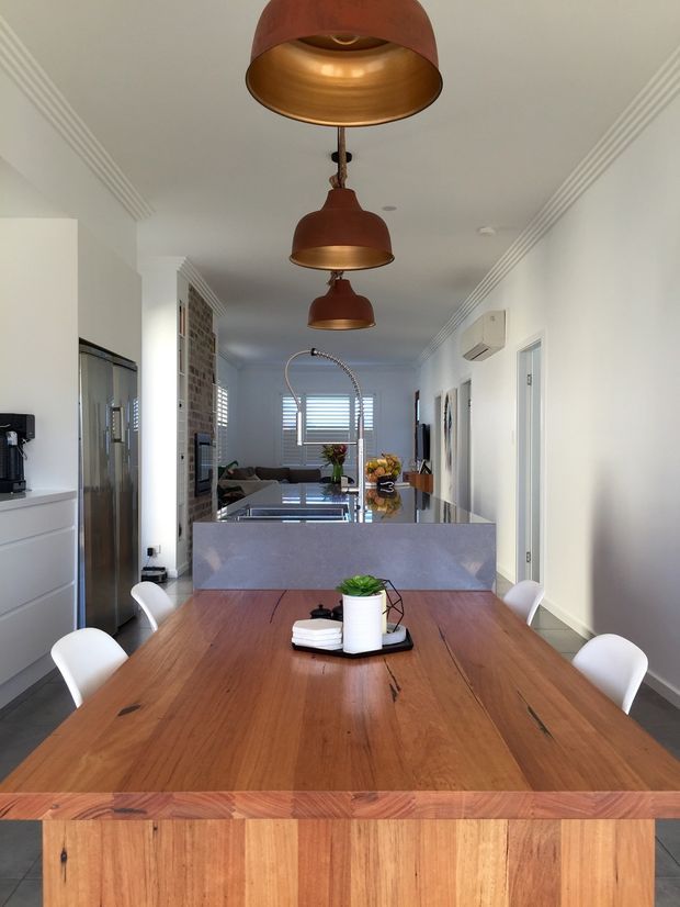 Bright open-plan interior viewed from the dining area toward a contemporary kitchen and living space. A large solid timber dining table with white chairs occupies the foreground, with a stone island and dark reflective benchtop beyond. Three copper pendant lights hang in a row beneath ornate ceiling cornices. White cabinetry, stainless-steel refrigeration, exposed brick, timber flooring and plantation shutters create a warm, refined residential setting.