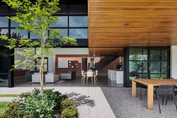 Contemporary residential courtyard viewed from outdoors, with a covered timber-lined terrace, large black-framed glazing, and an open-plan interior containing living, dining, and kitchen areas. The hardscaped courtyard includes a landscaped planter, outdoor dining table, and built-in-looking timber soffit and structure. The interior features a brick feature wall, dark stair, neutral furnishings, and abundant indoor-outdoor connection.