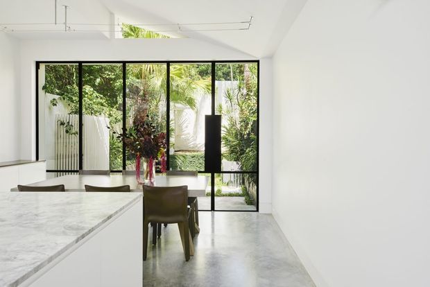 Bright contemporary open-plan dining and kitchen space with a long marble island in the foreground, a simple dining table and chairs, polished concrete flooring, white walls and ceiling, and full-height black-framed glazing opening toward a lush private garden courtyard. Minimal pendant lighting and clean-lined detailing create an airy indoor-outdoor connection.