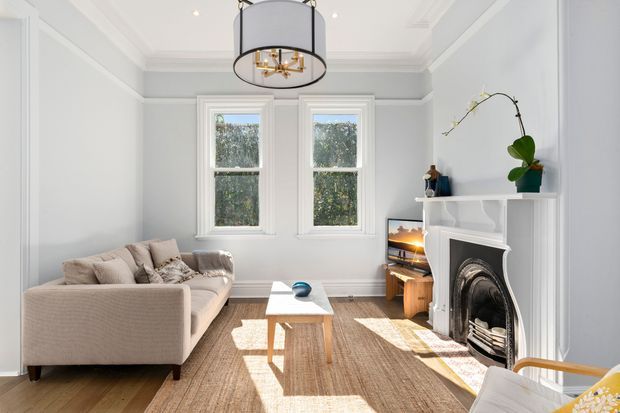 Bright, refined living room with pale blue-grey paneled walls, ornate ceiling mouldings, two sash windows, a white decorative fireplace, and a neutral sectional sofa arranged around a simple timber coffee table. A large drum pendant light, woven rug, timber flooring, and understated styling create a calm contemporary-classic interior.