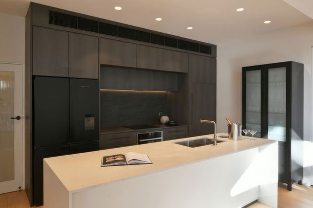 Contemporary kitchen featuring a large light-coloured island with integrated sink and tap, dark full-height cabinetry, a black refrigerator, dark splashback, under-cabinet lighting, and a tall black-framed glass display cabinet. Recessed ceiling lights and strong natural light create a clean, refined contrast between pale and dark finishes.