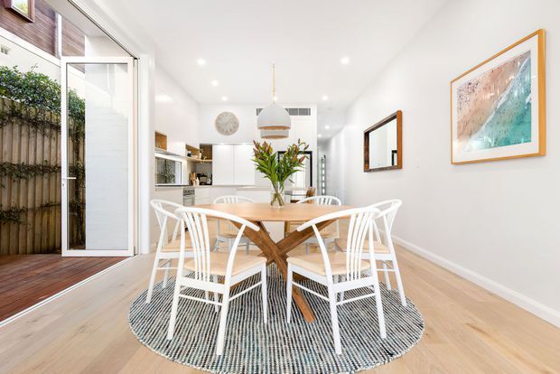 Bright open-plan dining area with a pale timber dining table and white spindle-back chairs arranged on a round textured rug. The room has white walls and ceiling, light timber flooring, a compact kitchen visible in the background, recessed downlights, a pendant over the table, framed artwork, and large glazed doors opening to a timber deck or courtyard.
