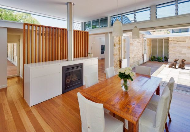 Bright open-plan dining and living area with a polished timber dining table, white upholstered dining chairs, a built-in fireplace within white cabinetry, and a vertical timber slat room divider. High-level louvred glazing and clerestory windows bring in abundant daylight, with a stone-paved courtyard visible beyond the large openings.