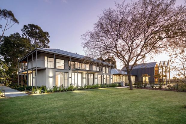 A contemporary two-storey residential property viewed from a landscaped lawn at dusk. The main house has a light rendered facade, extensive floor-to-ceiling glazing, slim dark metal framing, projecting upper-level balconies, and a low-pitched metal roof. A separate dark-clad gabled pavilion or outbuilding sits to the right, connected by a paved path and enclosed garden area. Mature trees and planting surround the expansive lawn.