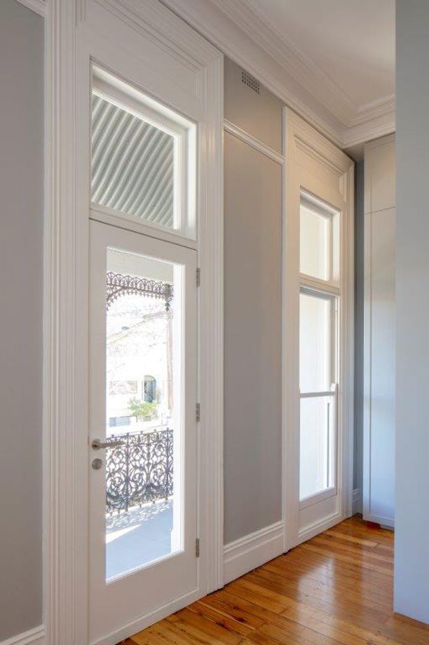 Bright, refined entry hall with a glazed white panel door, matching tall windows, detailed wall mouldings and wainscoting, a decorative plaster ceiling cornice, and polished warm timber floorboards. Ornate balcony railings are visible through the door glazing.