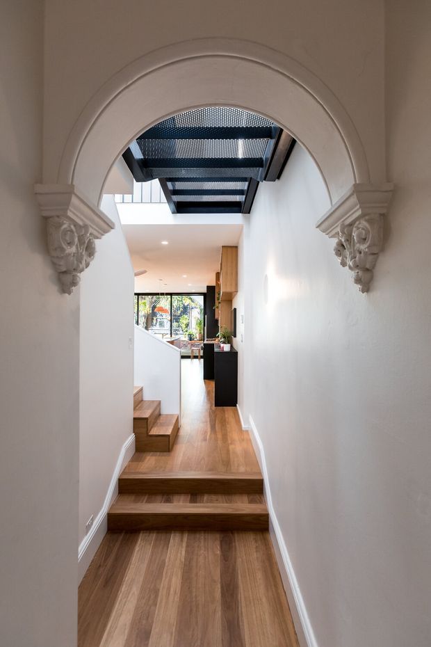 A narrow interior hallway viewed through a decorative arched opening with ornate plaster corbels and moulding. The corridor has warm timber flooring and steps leading toward a bright open-plan living area, with a contemporary black perforated skylight or stair void overhead and glimpses of built-in cabinetry and glazing beyond.