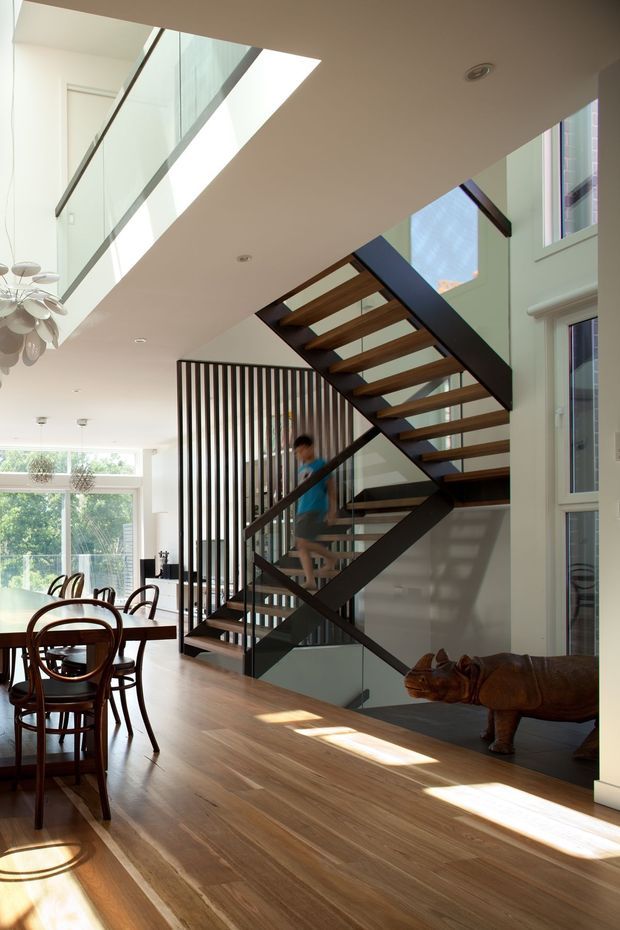 Bright open-plan residential interior featuring a sculptural floating staircase with dark metal stringers, timber treads, and glass balustrades. A vertical black slat screen separates the stair from the dining area, which has a timber dining table and bentwood chairs. Large windows, a skylight, light walls, timber flooring, and a decorative rhinoceros sculpture create an airy contemporary setting.