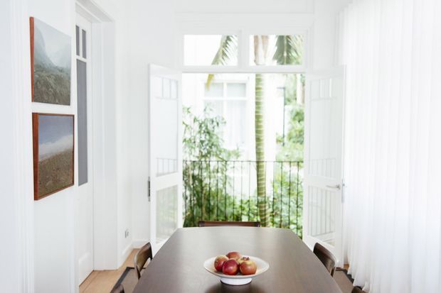Bright, airy dining room with a dark rectangular dining table and fruit bowl, surrounded by white walls, tall sheer curtains, framed artwork, and open glazed French doors leading to a leafy balcony or garden view. The space has abundant natural light and a clean, refined coastal feel.