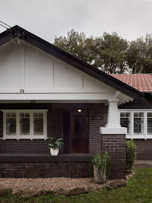 Front exterior view of a single-storey brick bungalow with a prominent gabled roof, dark brick walls, white timber-framed windows with shutters, and a covered entry porch. The facade includes decorative white gable detailing, a substantial porch column, dark roof trim, and a tiled roof section extending to the right. Low landscaping and potted plants sit in front of the house.