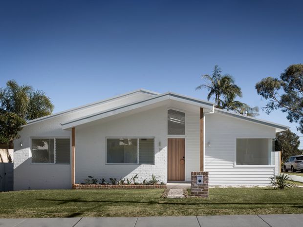 Front exterior view of a single-storey suburban home with a low-pitched asymmetric roof, white painted brick and horizontal weatherboard cladding, timber entry door, large windows, and a small landscaped front lawn.
