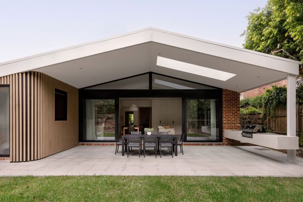 A contemporary single-storey rear extension opening onto a paved outdoor entertaining area. The structure has a broad flat roof with a long skylight, dark-framed floor-to-ceiling glazing, a timber-look vertical batten wall, exposed red brickwork, and a built-in outdoor barbecue bench. A dining setting sits beneath the covered terrace, with the interior visible through the glazed doors.