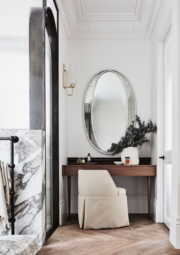 Elegant bright interior vignette featuring a compact timber console desk and upholstered chair beneath a large oval mirror. White walls and ornate decorative ceiling mouldings create a refined classic setting, while marble wall cladding, dark fluted paneling, brass wall light, herringbone timber flooring, and curated accessories add contrast and texture.