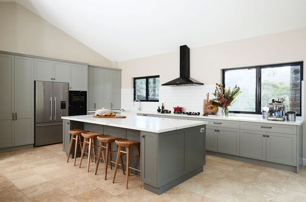 A spacious contemporary kitchen with a large central island, integrated seating for four, grey-green shaker-style cabinetry, white benchtops, stainless-steel appliances, a black rangehood, and large windows overlooking greenery. The room has pale tiled flooring and a bright, refined atmosphere.