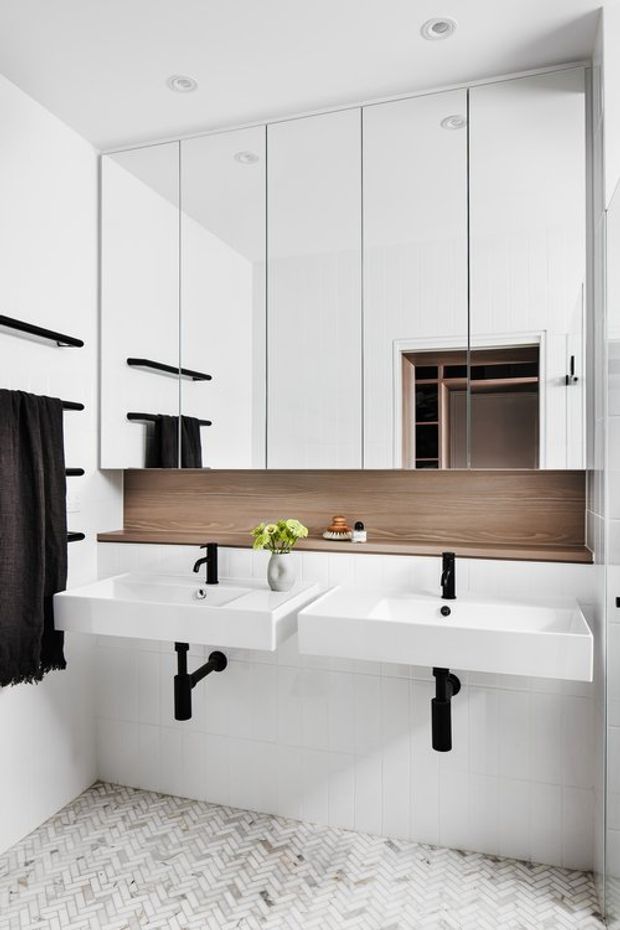 Bright contemporary bathroom featuring a double wall-mounted vanity with white rectangular basins, matte black taps and exposed bottle traps, a large mirrored cabinet, warm timber-look backsplash shelf, white tiled walls, and a white herringbone tile floor. Black towel rails and towels provide contrast.