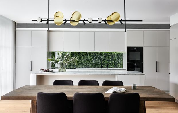 A bright, refined open-plan kitchen and dining area. The kitchen features full-height white cabinetry, integrated black appliances, a pale stone benchtop, and a large horizontal window overlooking greenery. A substantial dark timber dining table with black upholstered chairs occupies the foreground, beneath a sculptural linear pendant light with brass-gold spherical elements.