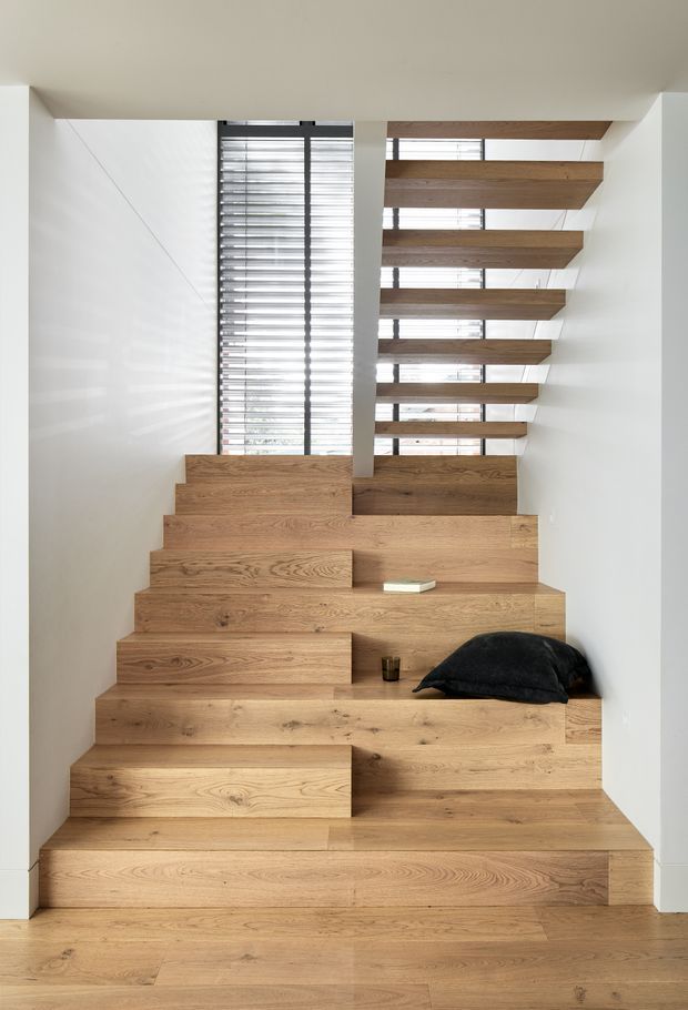 A minimalist internal stairway formed from broad, stepped timber treads and integrated seating or landing platforms. The stair rises beside a tall glazed opening with horizontal blinds, white walls, and exposed timber ceiling elements, creating a bright, sculptural circulation space.