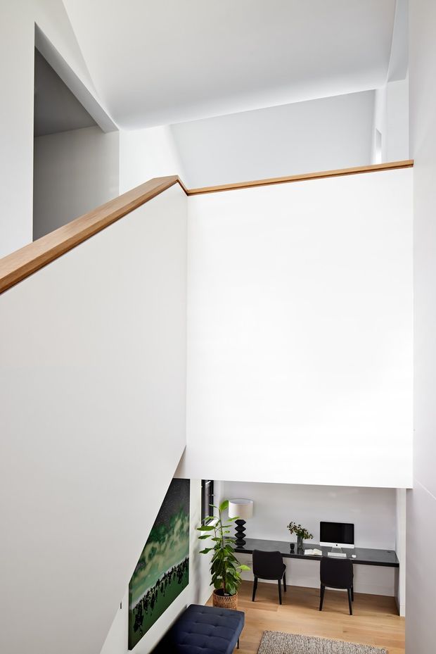 A bright, minimalist stairway and landing with crisp white walls and ceiling, a simple timber handrail, and a view down to a compact home office. The office includes a long built-in desk, two dark chairs, a computer monitor, indoor plants, timber flooring, and a large artwork.