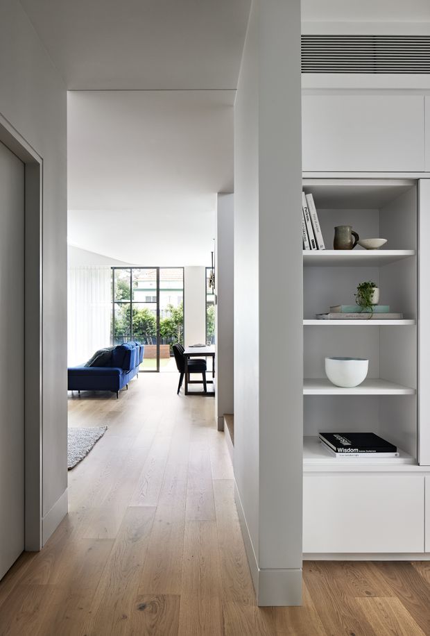 A bright, minimalist residential interior viewed from a hallway, looking toward an open-plan living and dining area. Pale timber flooring runs throughout, with white walls, a built-in white shelving unit displaying books and ceramics, a deep blue sofa, and full-height glazing opening toward greenery outside. Clean-lined cabinetry, concealed air-conditioning ventilation, and restrained furnishings create a calm contemporary atmosphere.