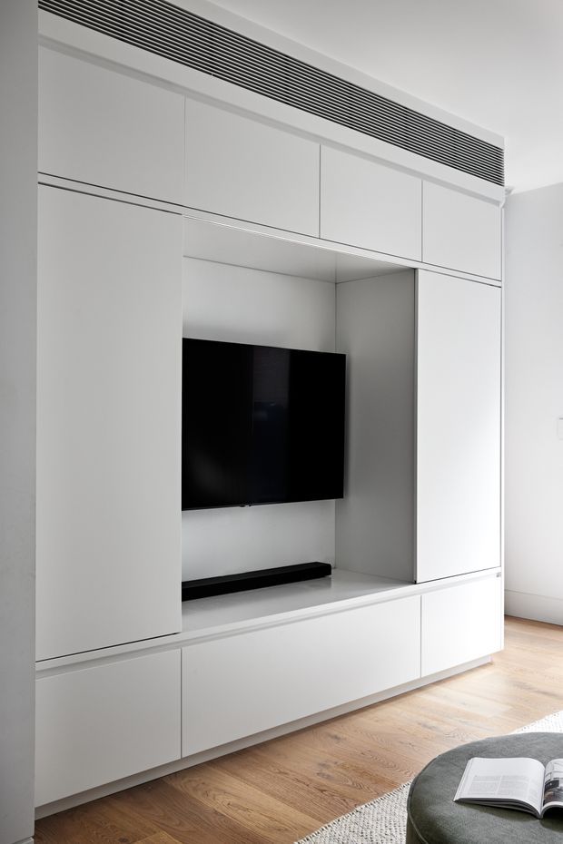 A minimalist built-in white media wall with extensive floor-to-ceiling cabinetry, a recessed television niche, concealed storage, and a long linear ventilation grille above. The room has pale timber flooring, bright white walls, and a partial rounded grey upholstered seat with an open book in the foreground.