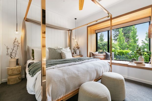 Bright, warmly styled bedroom featuring a four-poster timber bed with white and sage-green layered bedding, a large mirrored wardrobe or wall panel, sculptural pendant lighting, curved upholstered ottomans, and a generous timber-framed picture window overlooking greenery. The room has white vertical wall panelling, dark carpet, and a relaxed natural-material palette.