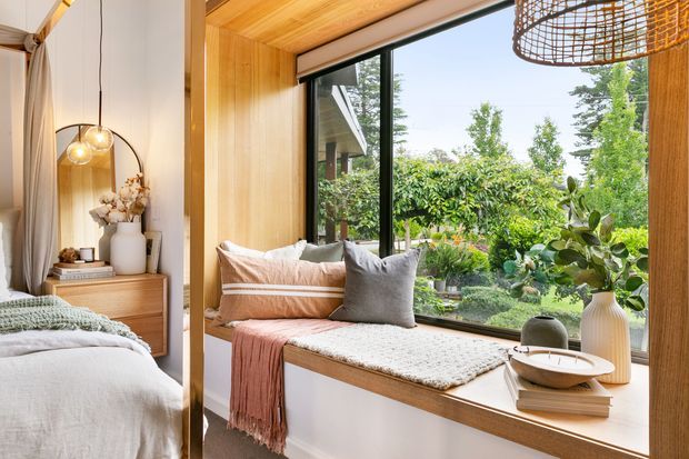 Warm, inviting bedroom with a built-in timber window seat overlooking a leafy garden. The room features timber wall and ceiling lining, a large black-framed window, soft neutral bedding and cushions, a woven pendant light, and a small timber bedside unit with decorative styling.